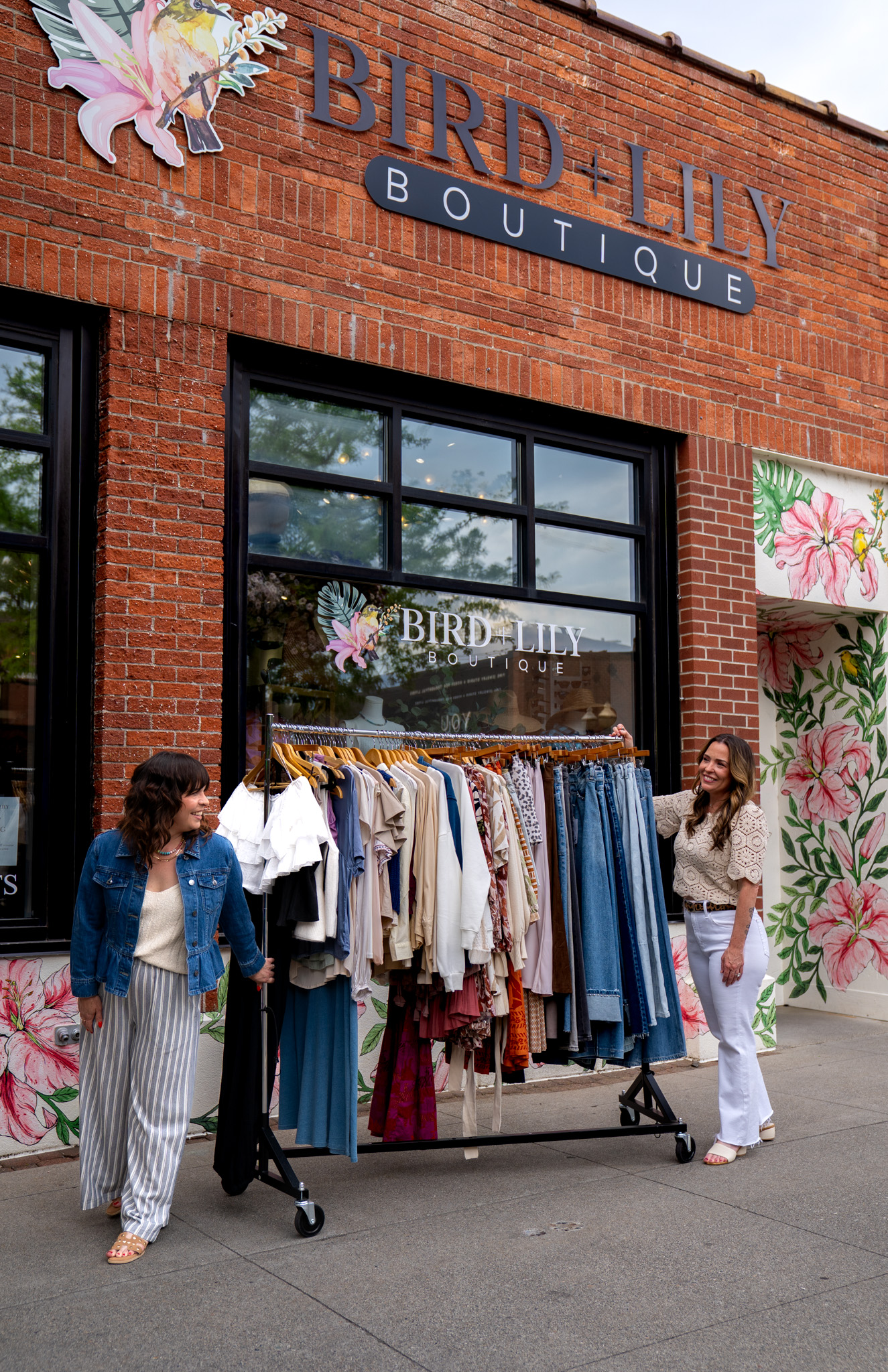Two women stand outside Bird + Lily Boutique in Coeur d’Alene, Idaho, beside a clothing rack displaying tops, dresses, and denim in front of the boutique’s brick storefront and floral mural.