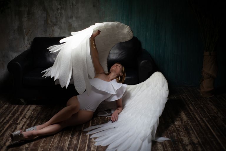 Full body gorgeous fit female wearing white bodysuit and big angel wings lying graciously on floor near leather couch
