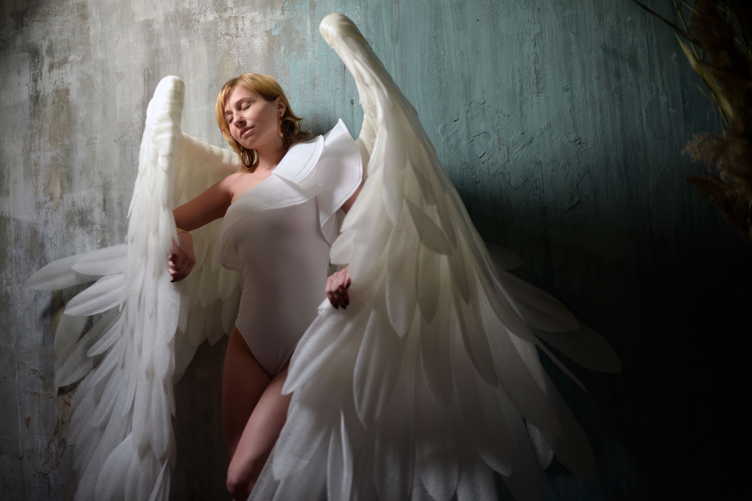 Young mindful female with closed eyes and fabric wings in white bodysuit against weathered wall